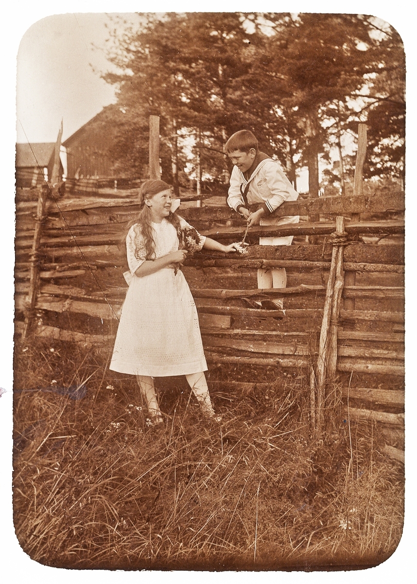 En flicka och en pojke vid en gärdesgård. Flickan har en liten blombukett i ena handen. På baksidan av fotot (tillverkat som vykort) finns följande text: "Fröken Elsa gratuleras hjärtligt av vännerna Sune och Margit".