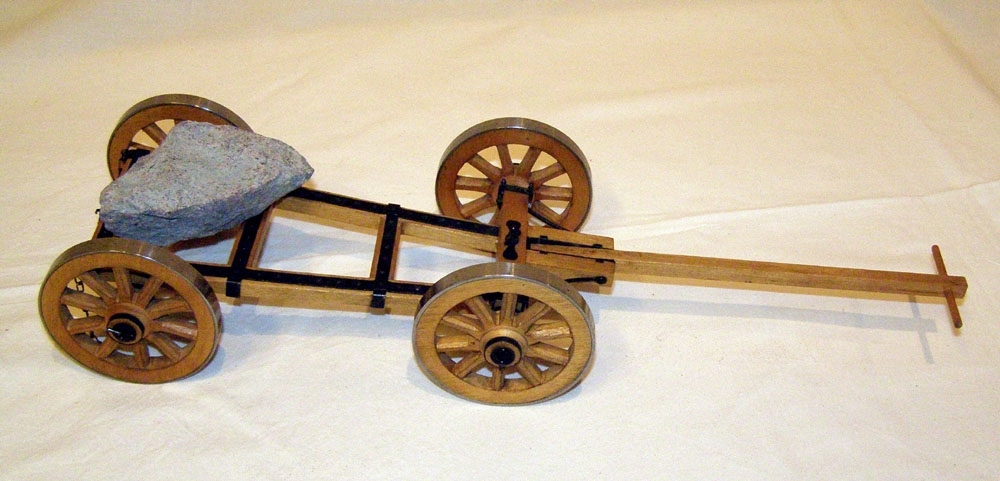 Modell i skala 1:10 av stenkärra med fyra hjul.
Modellen är tillverkad av trä med järnskoning. Axlar av järn. Järnskodda hjul.