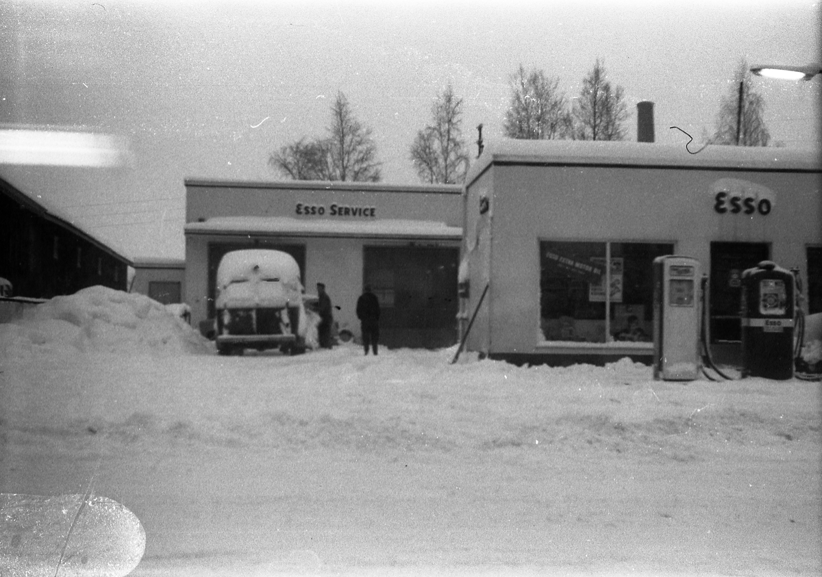 Snørikt gatebilde fra Lena, muligens vinteren 1962/63. Bildet er tatt gjennom vinduet i Lena Foto & Radio i "Raubua", i retning Esso bensinstasjonen. Foran smørehallen står en delvis nedsnødd buss.