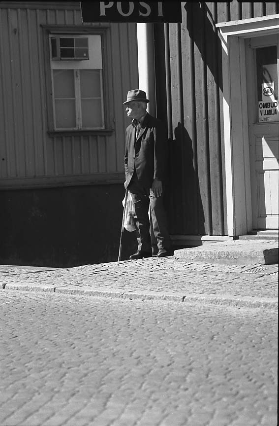 En äldre man, Karl Wristel med käpp, hatt och en kasse i handen, står vid husknuten och hörnet Brahegatan-Ribbagårdsgränd.
