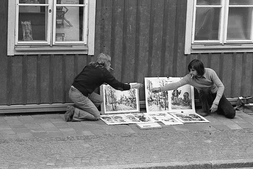 Två unga personer, med cigarett i munnen, sitter på huk vid en husvägg och säljer oramad konst. De sträcker sig mot varandran och lämnar möjligen över tändstickor/cigarettändare.