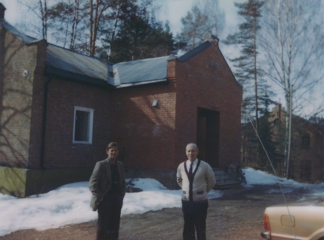 To menn foran teglsteinbygning som var kontor- messebygningen ved A/S Kistefos Træsliberi. Større fabrikkbygning i teglstein, tresliperiet, i bakgrunnen. Det er vinter og snø.
