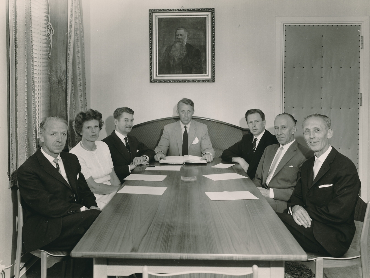 En kvinne og seks menn sittende ved møtebord med papirer foran seg. Innrammet portrettmaleri av Konsul Anders Sveaas henger på veggen.