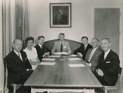 En kvinne og seks menn sittende ved møtebord med papirer for