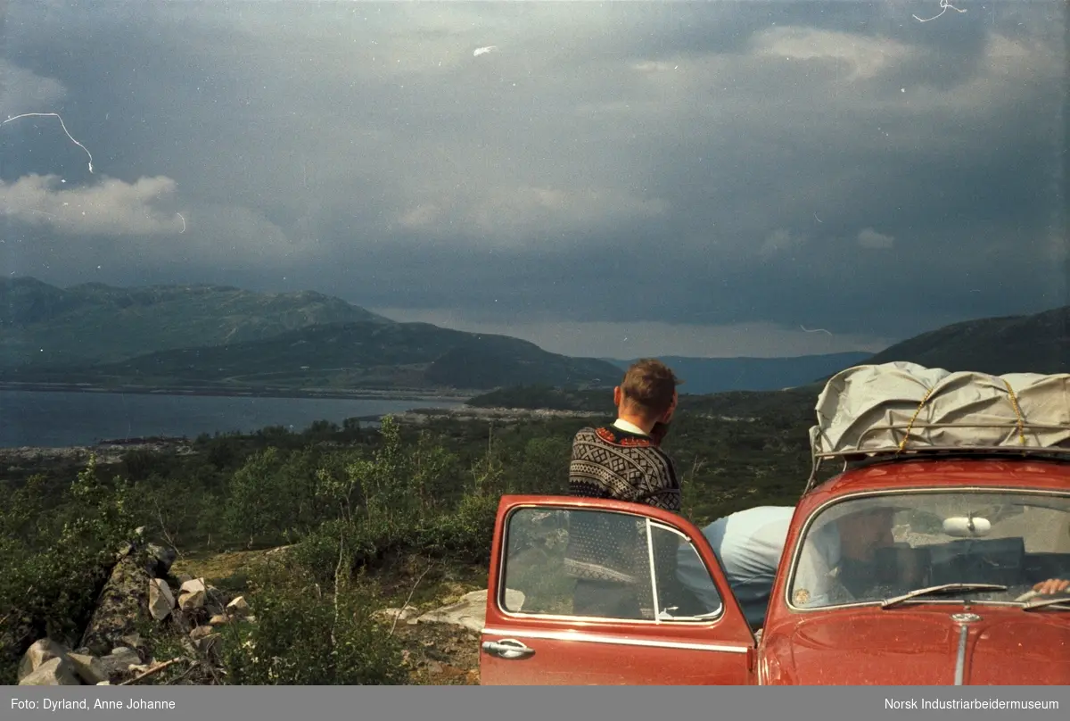 Tur til Røros. Olav O. Vågen i lusekofte står med ryggen til ved bildør og Trygve Vågen lener seg inn i en rød bil. Utsikt mot innsjø og fjell