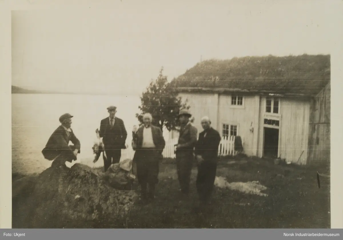 Fem menn på gårdstunet ved våningshuset på gården Skinnarland på Møsstrond. I bakgrunn innsjøen Møsvatn.
