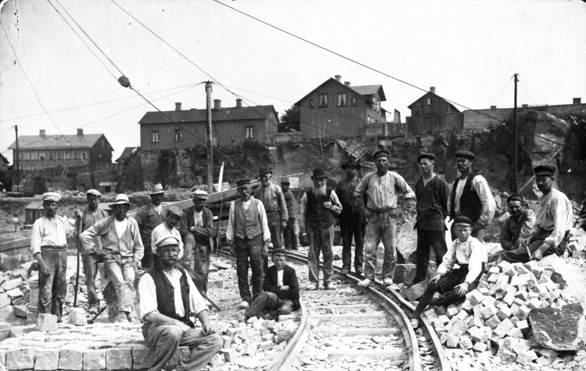 Stenbrottet vid Platsarna. Varbergs första fackförening. Ett stort antal stenarbetare, även två pojkar, kring rälsen och bebyggelsen på Platsarna i bakgrunden och huggen gatsten i förgrunden.