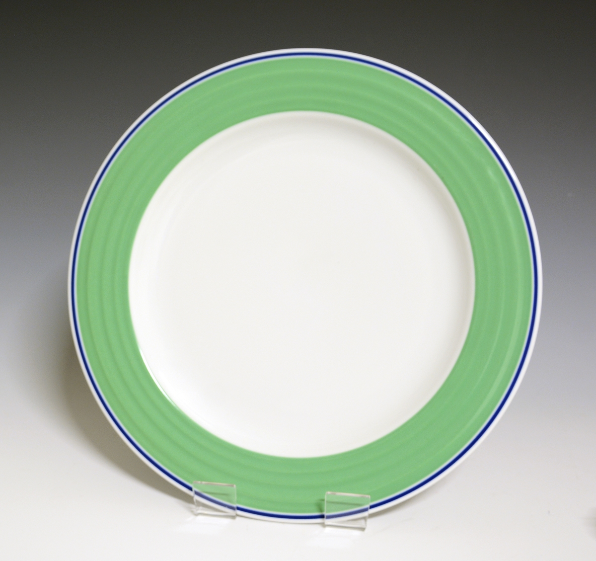 Middagstallerken av porselen med hvit glasur. Fanen dekorert med et grønt bånd med blå stripe ut mot kanten. I fanens gods riller. 
Modell: Saturn
Dekor: Saturn Grønn