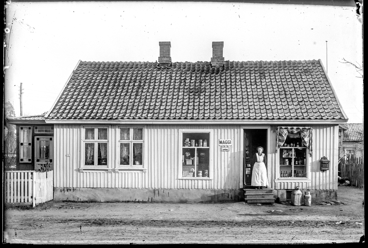 Klostergata 44 b i Moss. Butikk med reklame for Maggi og Sunlight.  Ukjent kvinne i døra. 
Bak til høyre er huset på MBIF.2000-014-118, Klommestensbakken 3.