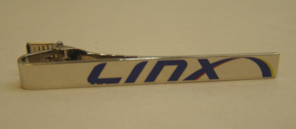 Slipsnål av silverfärgad metall, med namnet Linx i blått.
På baksidan en klämma som är räfflad på insidan för att hålla slipsen på plats.