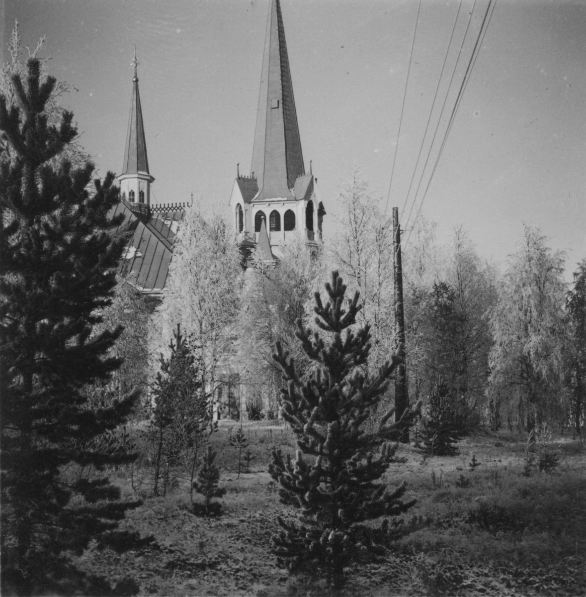 Exteriör av Jokkmokks kyrka, vintern 1942. Med träd och elledning i förgrunden.