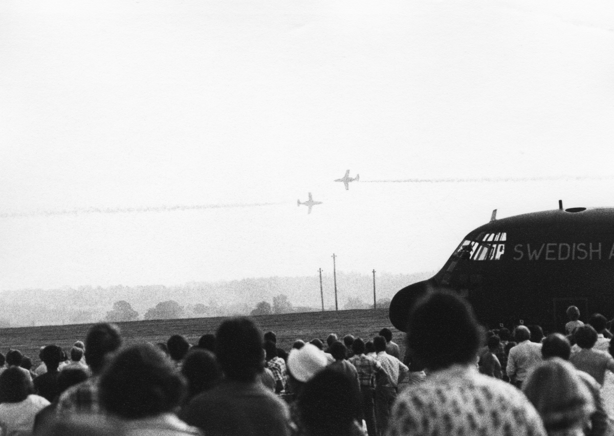 Avancerad flygning med två flygplan SK 60 ur Flygvapnets uppvisningsgrupp vid flygdagen på Malmen den 28 augusti 1976. Vid firande av Flygvapnet 50 år. Flygplanen syns på långt håll. I förgrunden syns publik samt del av flygplan TP Hercules.