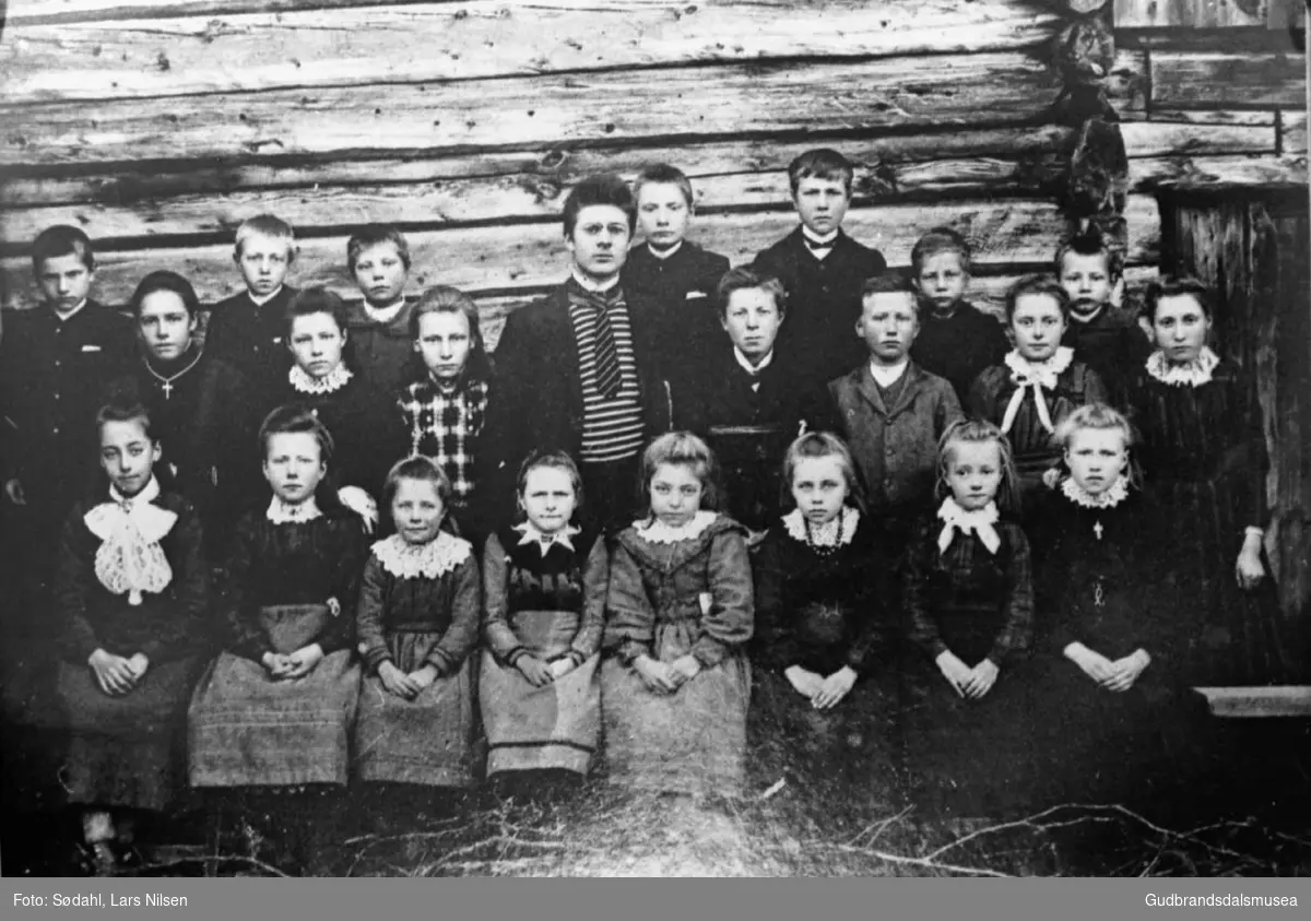 Gruppebilde, stor gruppe av skolebarn og en voksen samlet i husveggen i Uppistugu Bjølverud.

