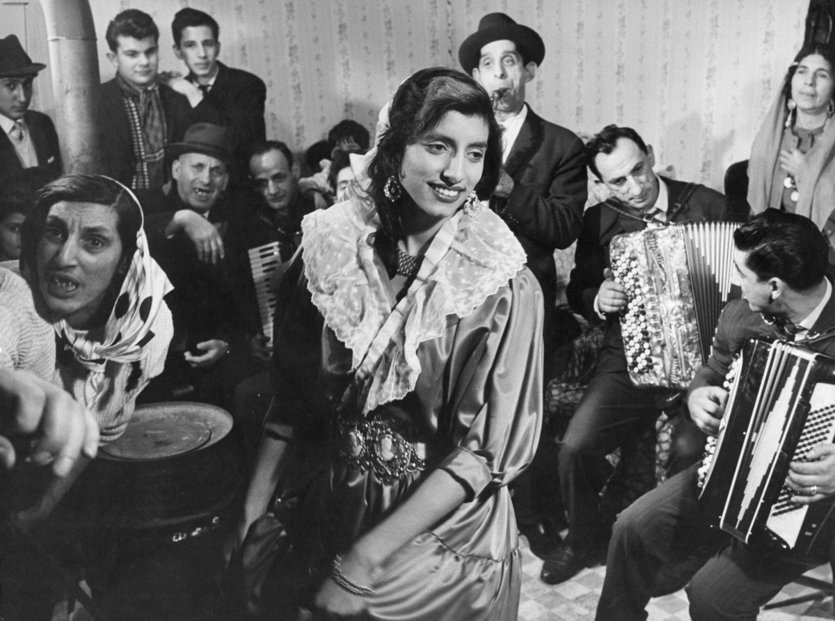 Inne i en bostadsbarack spelar en grupp romer musik. De har samlats runt en värmekälla bestående av ett oljefat med skorsten.