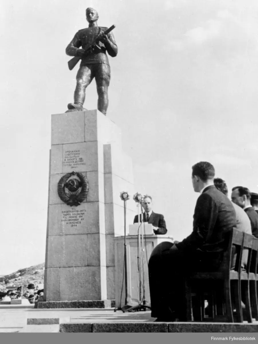 Avduking av russemonumentet i Kirkenes, 8.juni 1952. Monumentet er reist til Sovjetsamveldets tapre soldater til minne om frigjøringen av Kirkenes i 1944. En mann står nedenfor monumentet og holder tale, flere menn sitter på stoler like ved.