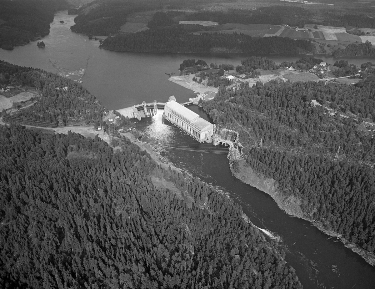 Solbergfoss kraftverk i Nedre Glomma, fotografert fra et fly som på opptakstidspunktet befant seg på Spydeberg-sida av elva, vest for kraftverket. Sentralt i bildeflata ser vi kraftstasjonen og dammen, bygd i perioden 1917-1924. På dammen, til venstre i den øvre delen av bildet, ser vi påstikkingslensa, som samlet tømmer som skulle føres forbi anlegget via ei tømmerrenne som dels var sprengt inn i fjellet på Spydeberg-sida av elva. Vi skimter utløpet av renna som et parti med kvitskummende vann ved vestre elvebredd nederst til høyre i bildet. Kraftstasjonen lå på Askim-sida.