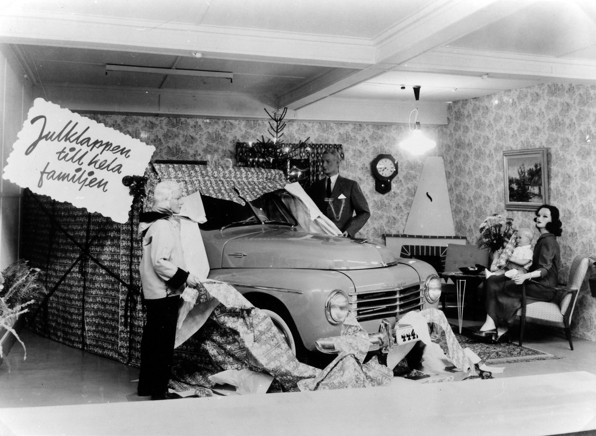 Alingsås Bil- och motorverkstads julskyltning i en, troligtvis hyrd lokal, i kv. Storken 5, ca 1955. Skyltodockor i en tidstypisk hemmamiljö, kvinna i fåtölj med barn i knäet, man som öppnat ett stort paket med inslagen volvobil och pojke som nyfiket tittar på från andra sidan.