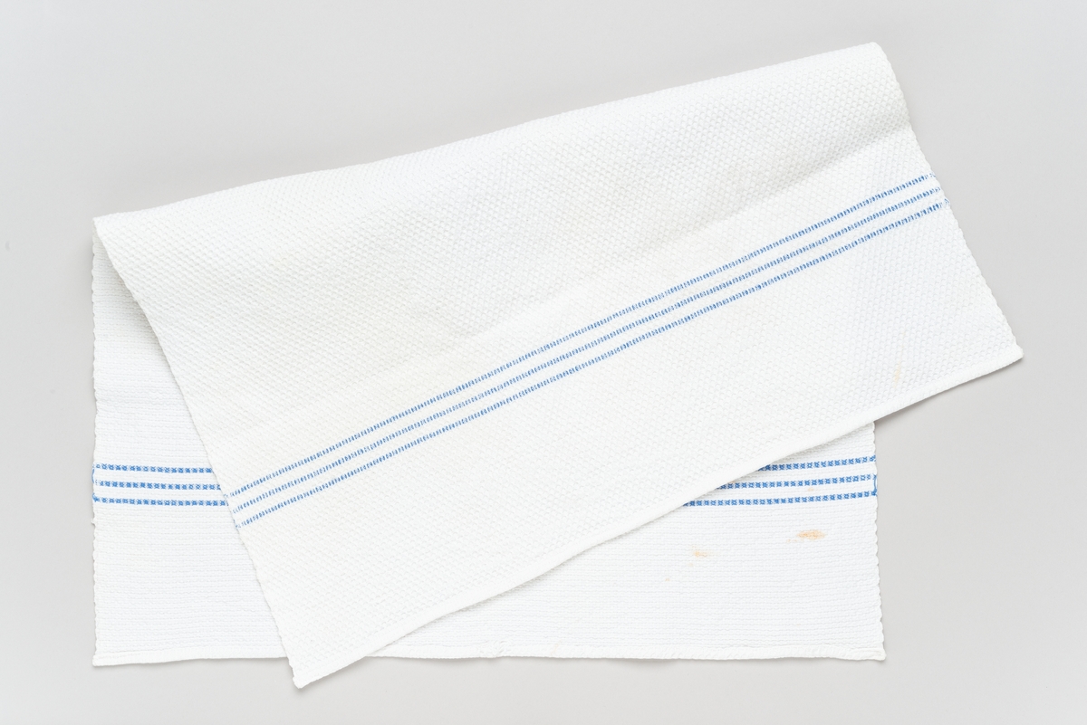 Rektangulært håndkle, hvitt med tre blå striper i hvert kortende. Den ene falden har revnet, kan være spor etter hempe.
