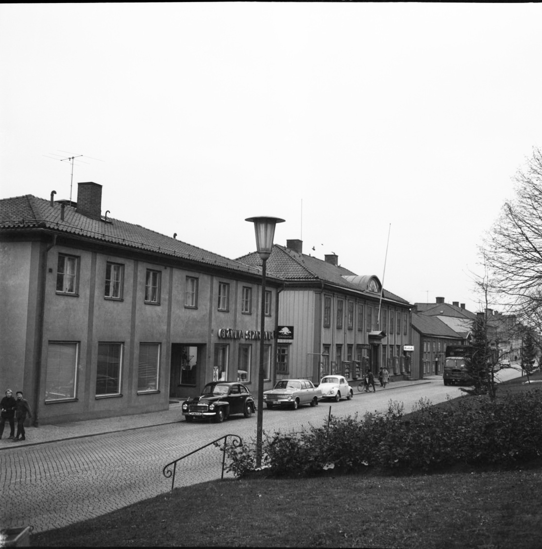 Brahegatan med Sparbankshuset och Hallska gården. Bilar står parkerade utmed fastigheterna. En lastbil kommer från höger och till vänster går två pojkar på trottoaren.
