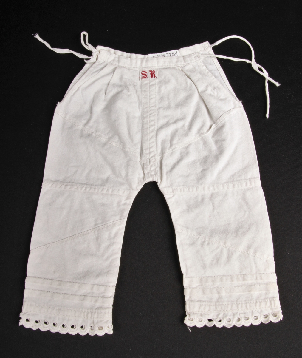 Hvit bomullsmamelukk til dukke eller spedbarn, med knyting i sidene og blondekant nederst på beina.