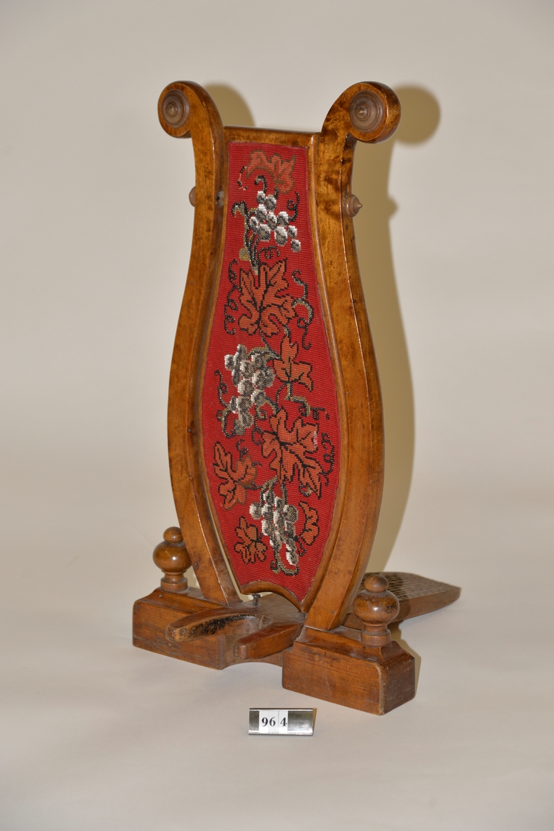 Stövelknekt bestående av en lyrformad träställning med en mittplatta av röd textil prydd av broderi.