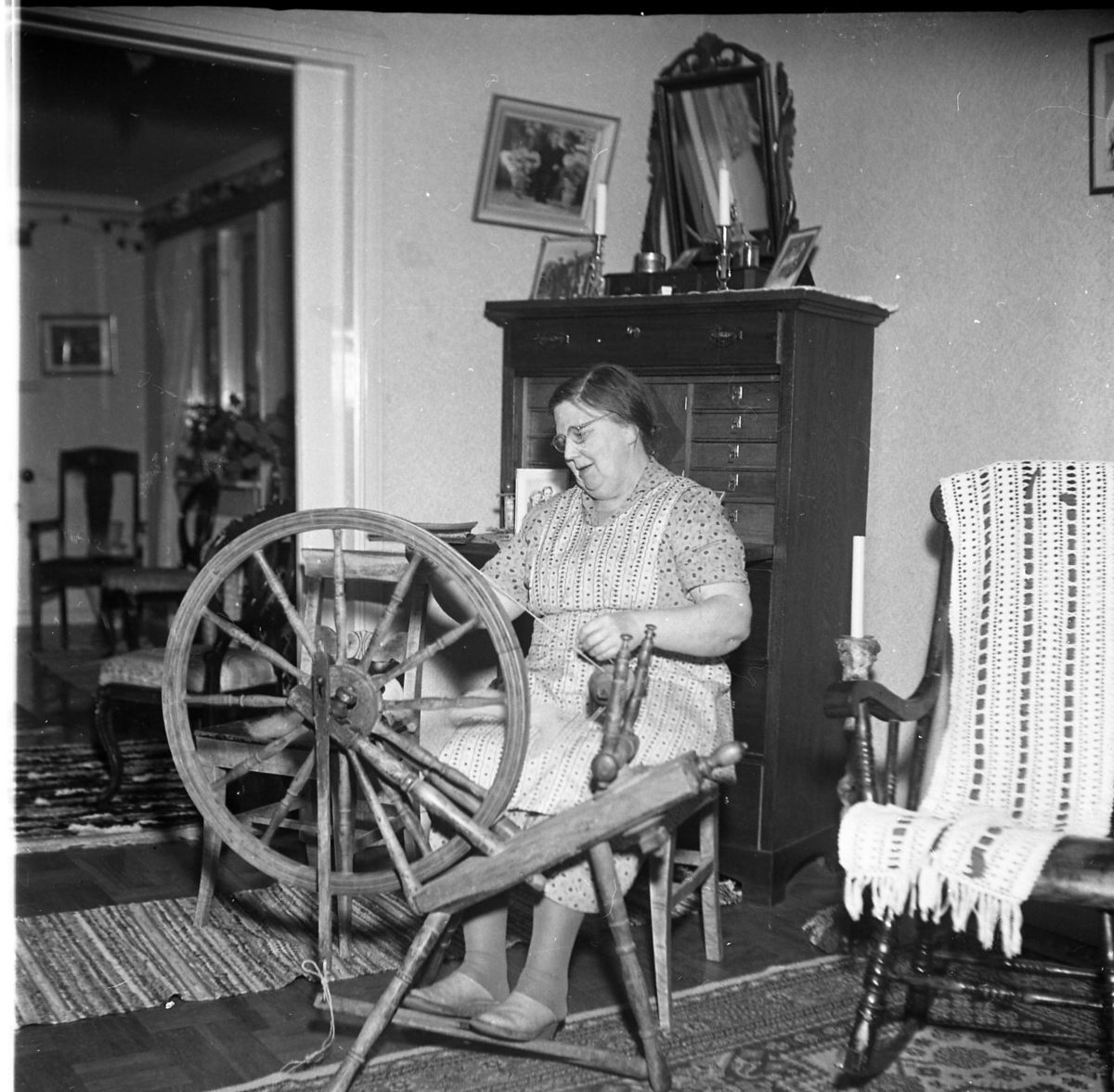 Fru Rut Sääv arbetar vid sin spinnrock i sitt hem. Bakom henne står en hög sekretär med en pigtittare ovanpå. Till höger en gungstol med troligen en virkad genombruten sjal