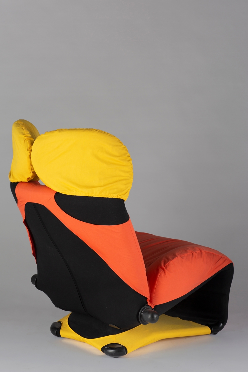 Svart stålramme med stor pute i skumgummi trukket i svart kunststoff. Oransje polyestertrekk i tillegg på sete og rygglen, og gult polyestertrekk på to hodeputer og sjeselongdel. Disse trekkene er løse, utskifts- og vaskbare. Rygglen og sete er bakoverlent, og i oppreist tilstand er liggedelen bøyd under setet. Ved hjelp av håndtak på hver side av stolen, kan den fleksibelt vippes ut til en sjeselong. Håndtakene justerer også ryggens og hodeputenes vinel.