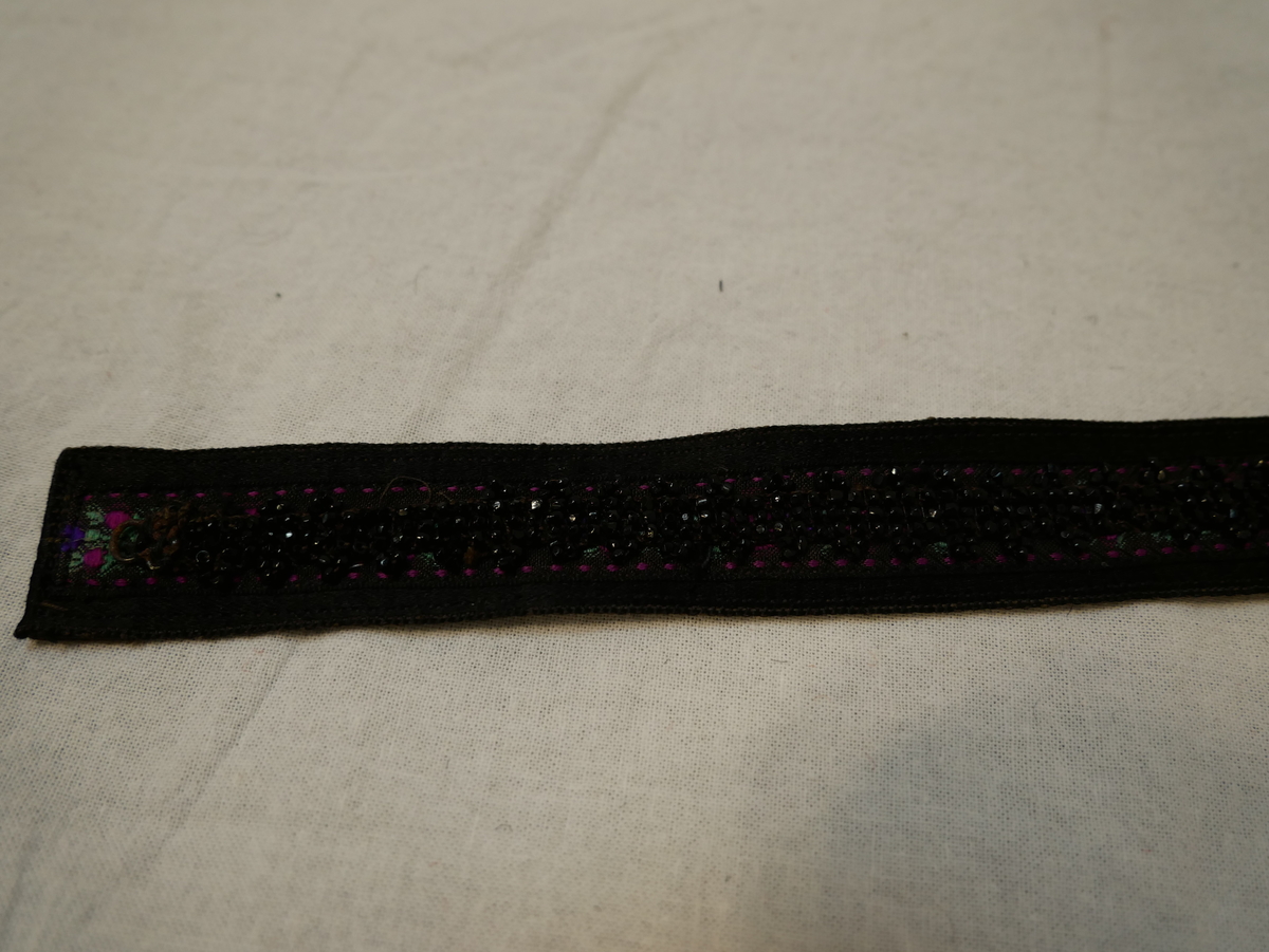 Svart silkeband med eit rosete silkeband sydd på langs midten. På silkebandet er set sydd fast eit band med svarte perler. Baksida fôra med brunt glansa lerret. Hekte i eine kortsida, krok i den andre.