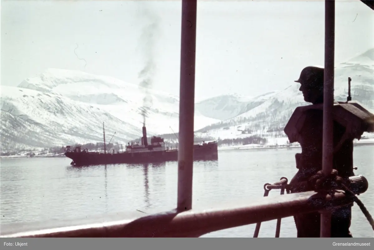 Tromsøysundet med Tromsdalstind i bakgrunnen. Skipet midt i bildet har påskrift NORGE. Navnet er vanskelig å lese. Soldat med våpen i forgrunnen til høyre.