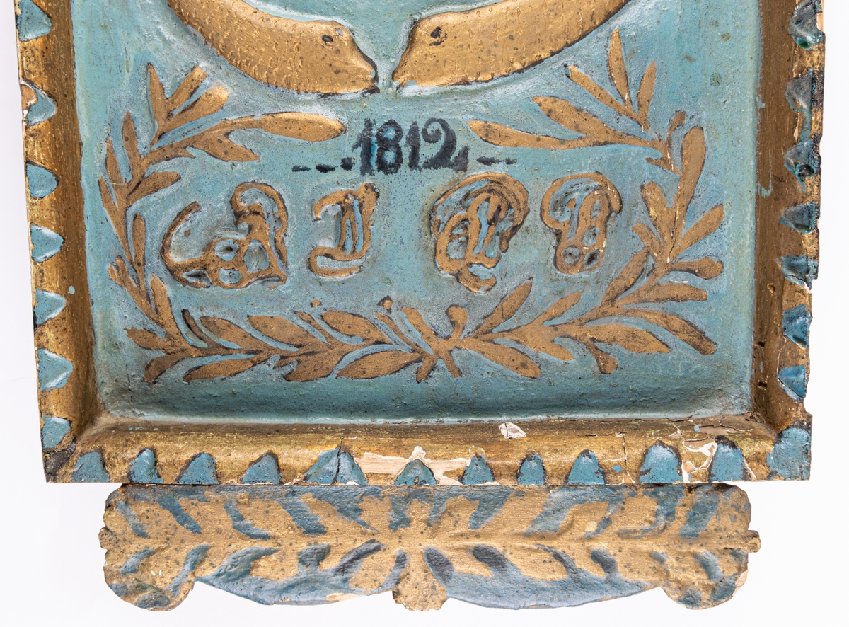 Klockfodral i skuret trä, blåmålat och förgyllt. Reliefbilder; krona, fiskar, delfiner, växtornament samt svårlästa bokstäver (Carl Johans namnschiffer?) samt märkt 1812. Runt hål i mitten.
