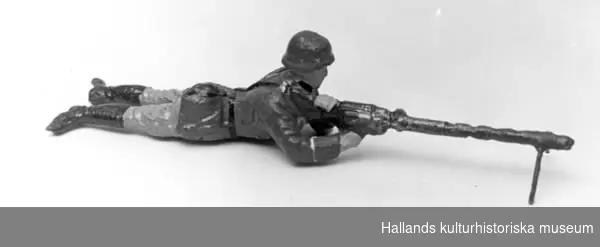 Leksakssoldat av trämassa på ståltrådsskelett i tysk uniform. Soldaten ligger raklång och skjuter med en kulspruta. Målad i grönt, brunt, svart, grått och skärt.