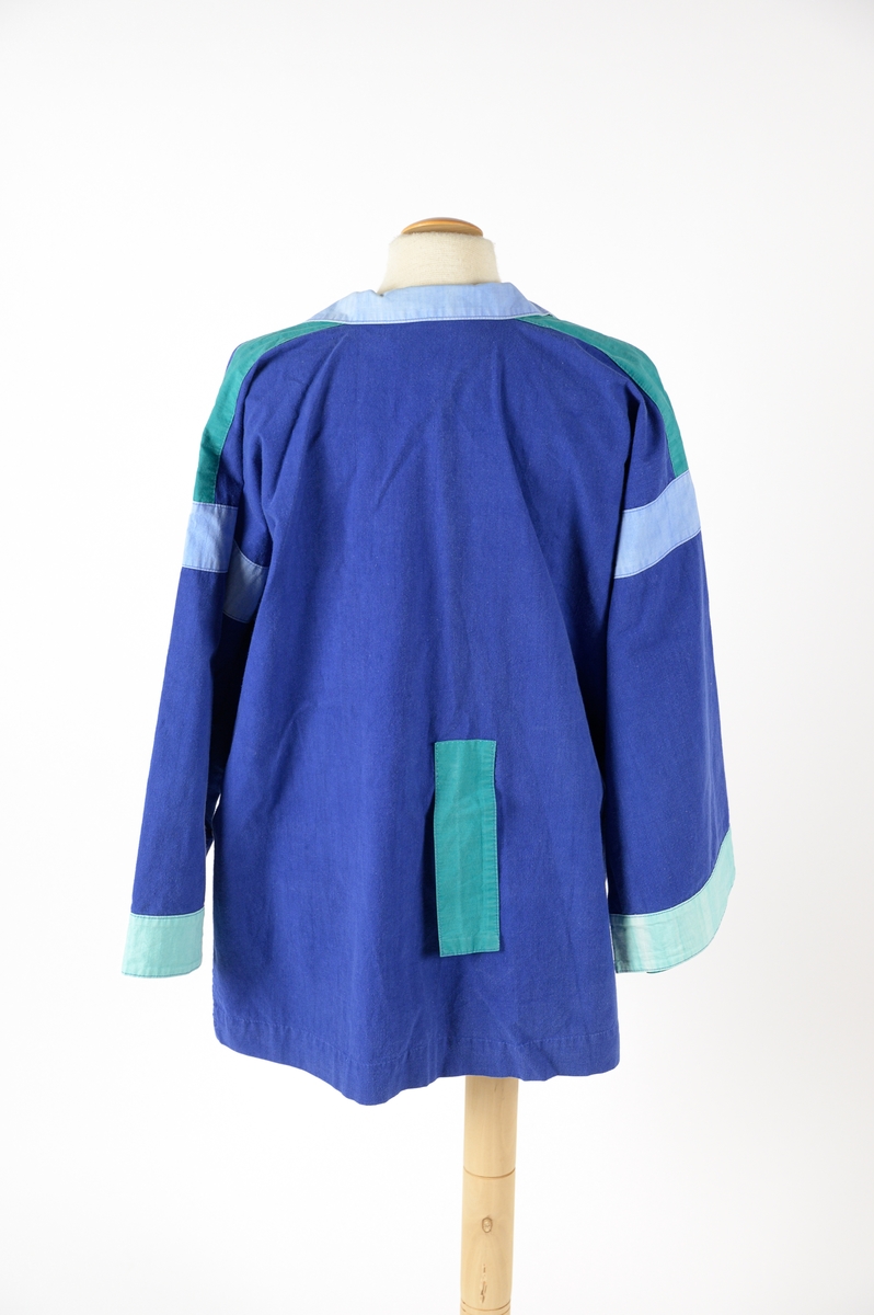 Skjorta designad av Barbro Sörman för Twilfit. Skjortan är kornblå med detaljer på ärmar, fickor och krage i ljusblått och mintgrön 100% bomull. Vid modell som dras över huvudet, krage och slag samt att det är sprund på båda sidor, två stora utanpåliggande fickor nedtill, dessa har en kant av ljusblått tyg. Gröna klaffar och ljusblå kilar vid ärmen, mintgrön bård vid ärmslut. Baktill sitter en grön rektangulär detalj som ev. sytts dit i efterhand för hand och den täcker ett litet hål. Skjortan har några fläckar på framsidan samt att den troligtvis är något blekt.