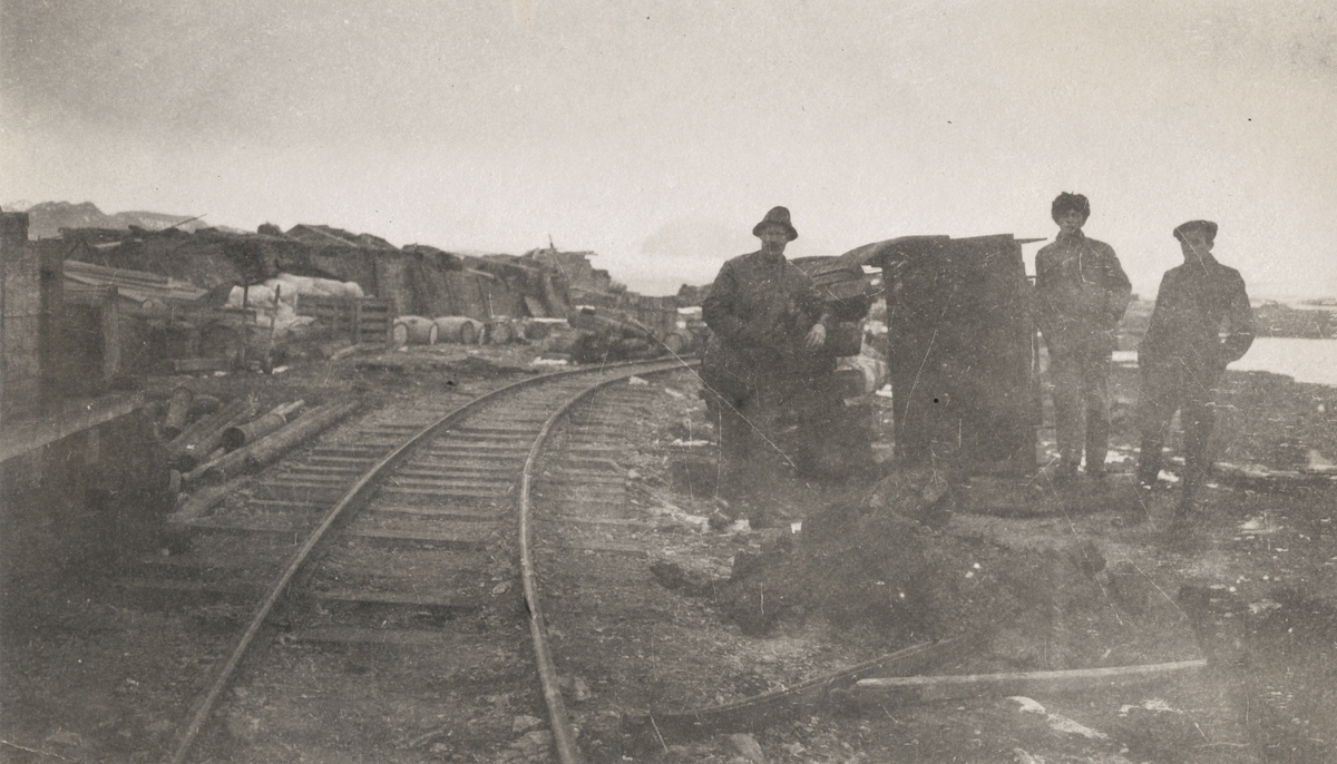 Motiv av tre menn som står ved jernbanen og en hel del materialer og gods for bygging av anlegg til kullgruvedrift.