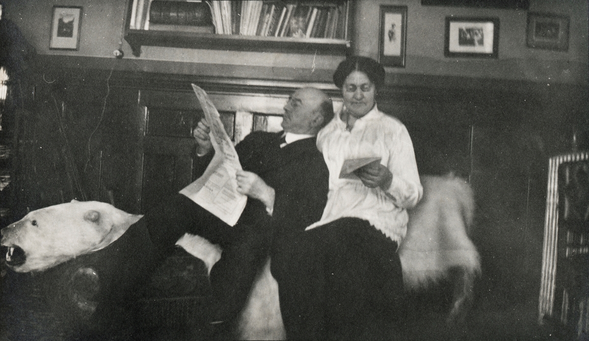 En mann og kvinne sitter på et isbjørnskinn i ei stue og leser.