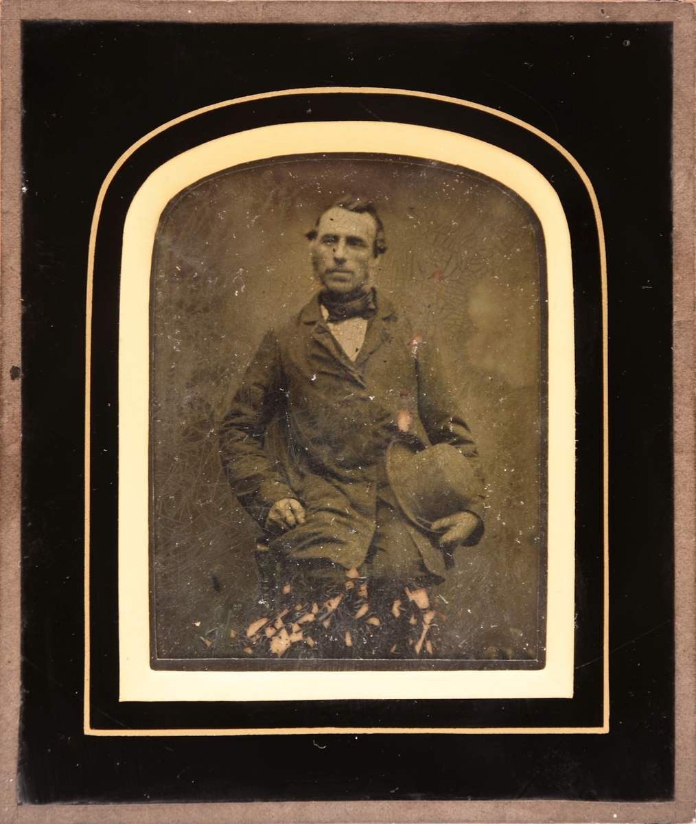 Portrett av mann i mørk dress med kalvekryss i halsen og hatten i hånden. Ambrotyp fototeknikk. Dette bildet er en glassplate der motivet er positivt fremkalt fra et glassplate-negativ. På baksiden en opphengsring.