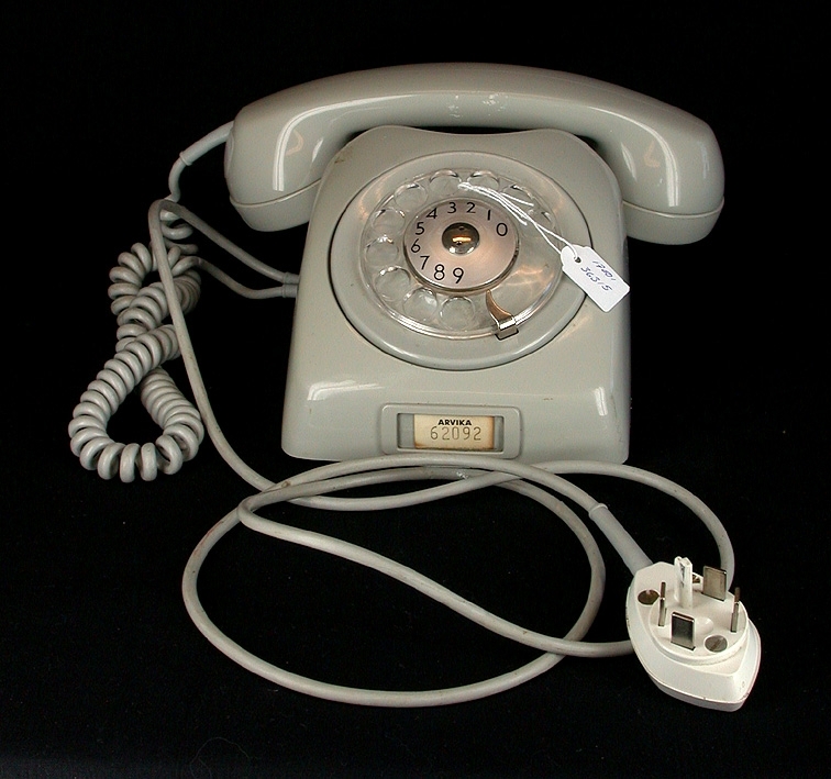 Grå telefon av modellen Dialog, tillverkad av LM Eriksson. På fronten liten plastförsedd insats att placera lapp om telefonnummer i. I denna insats sitter lapp med texten "ARVIKA 62092".