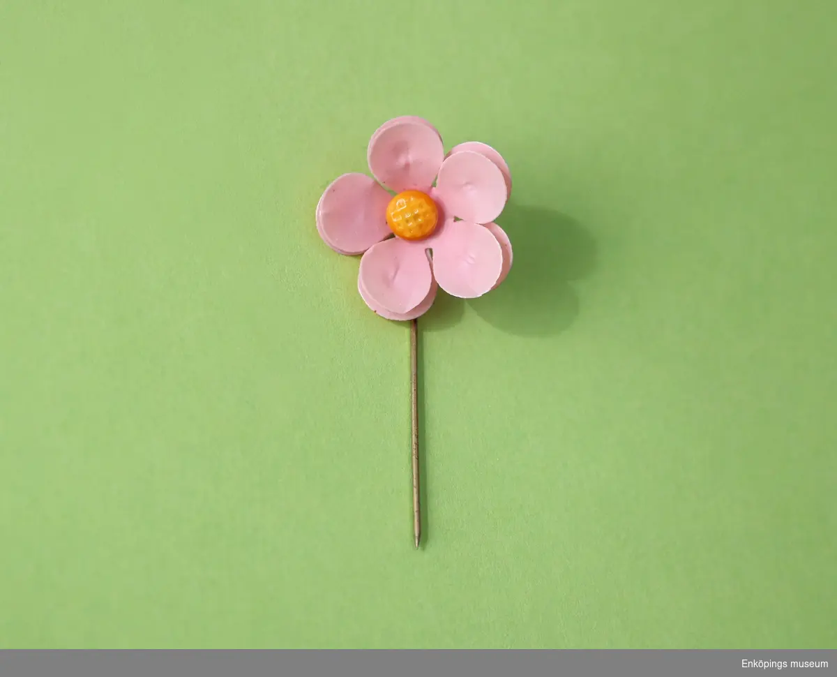Majblomma från år 1911.
Blomman är gjord av rosa celluloid och har fem blad i dubbla lager, totalt 10 blad och en gul mittknapp, även denna gjord av celluloid. 
Det som håller blomman samman är en nål av mässing.