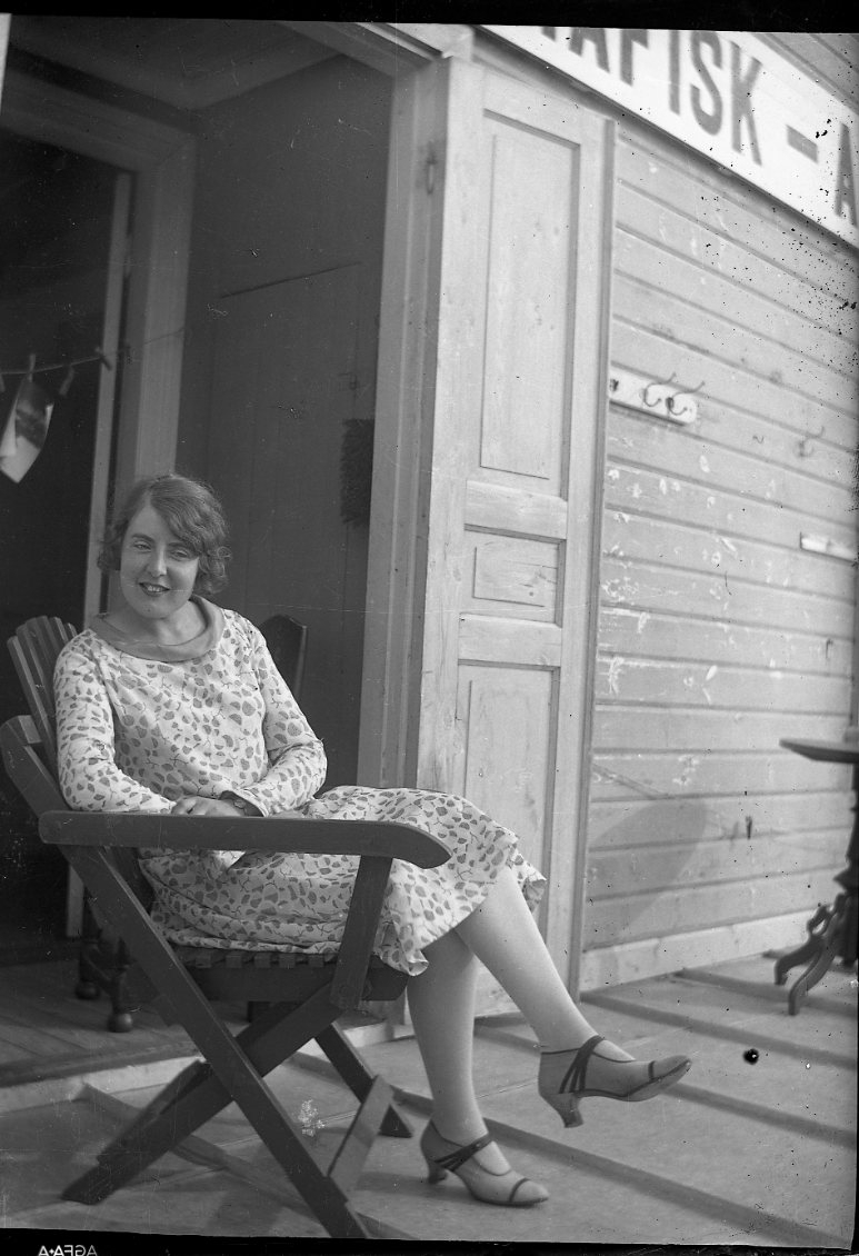 En kvinna i ljus mönstrad klänning sitter i en trädgårdsstol, troligen på balkongen till Calla Sundbecks fotoateljé i Gränna. I bilden överkant anas en skylt med texten: "...AFISK-A..."