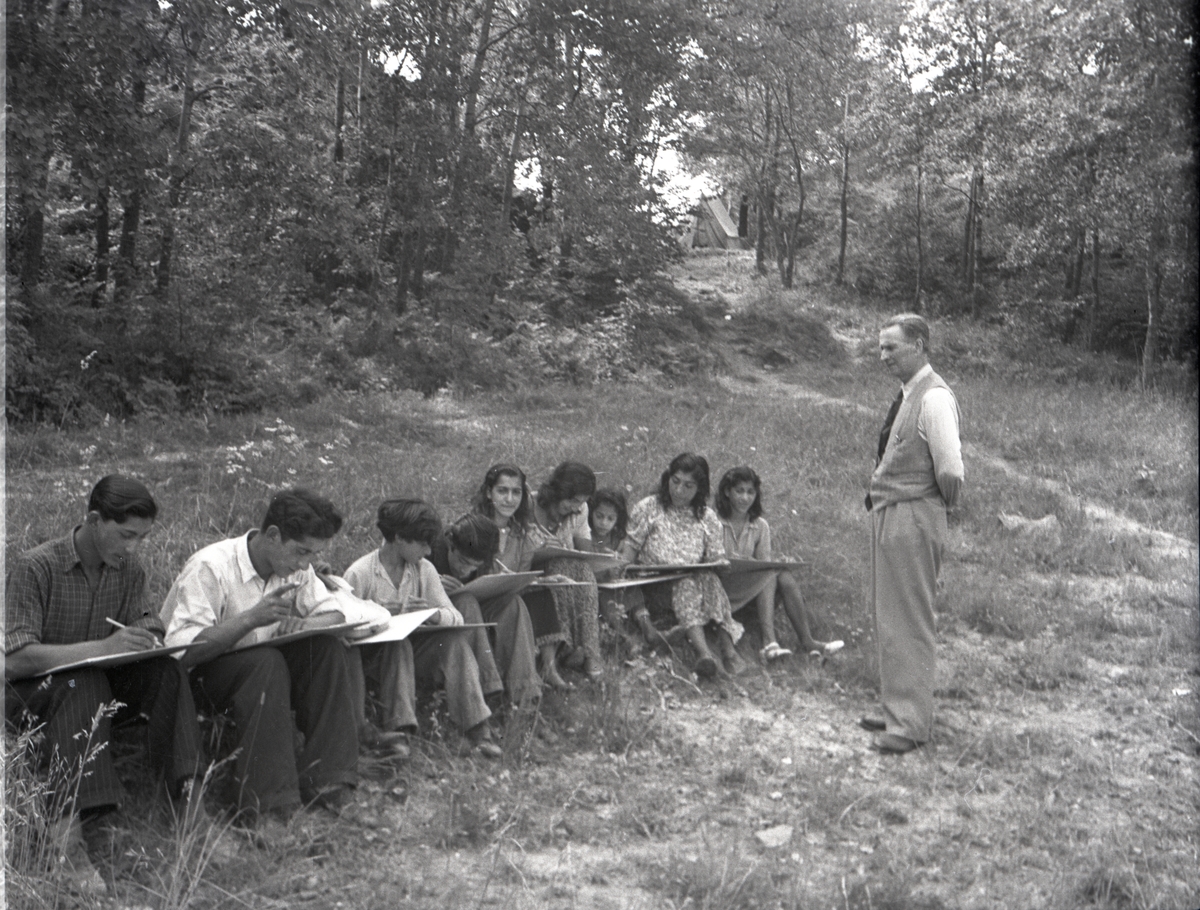Bilden är troligtvis tagen i samband med någon av de kurser som arrangerades för romer i mitten av 1900-talet, som kompensation för förlorad skolgång. Ett antal barn sitter på rad med skrivplattor i hand, intill dem står en lärare och ser på.