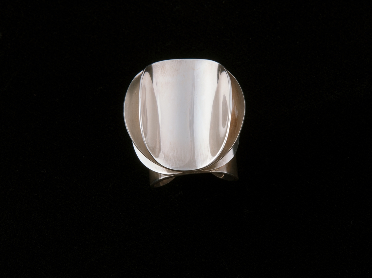Består av tre like store sirkulære sølvringer som er satt sammen langs en midtakse. På hver siden av aksen er ringene bøyd oppover i ulik grad, symmetrisk på hver side. Dekoren er festet på en fjerde rund form som danner ringen rundt fingeren.