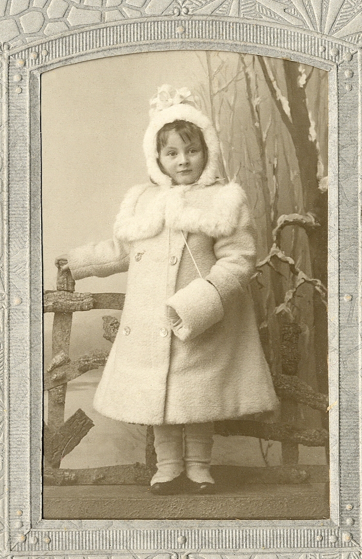 En liten flicka i ljus vinterkappa med svandunsgarnering och dito hätta. I ena handen håller hon en muff.
Helfigur. Ateljéfoto.

Fotografens dotter.