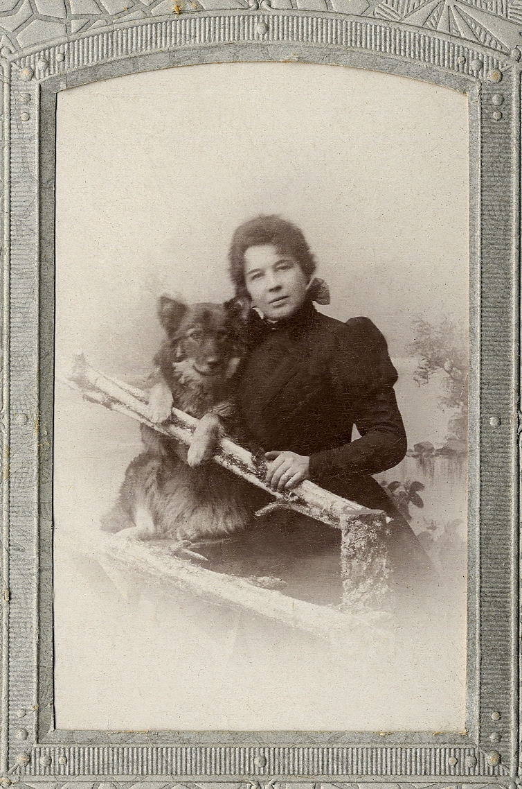 En kvinna i mörk klänning och hög krage tillsammans med en hund vid en "gärdesgård". 
Knäbild. Ateljéfoto.