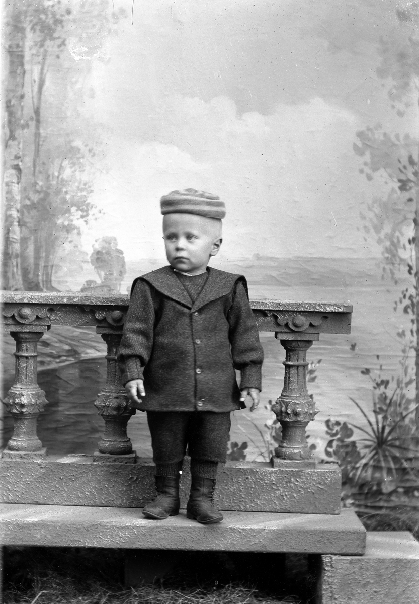 Liten gutt i dress og lue, står på steinhelle.