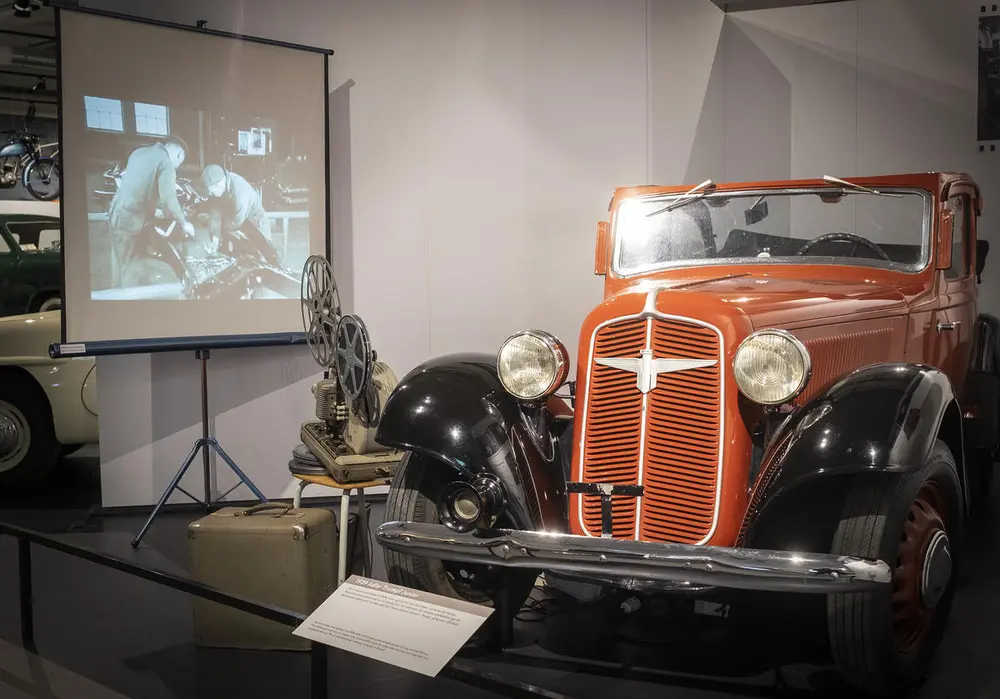 Bildet er fra Norsk kjøretøyhistorisk museum. Bildet viser en bil og et lerret som viser to arbeidere på en karosserifabrikk.