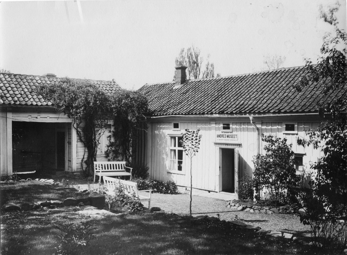 Gårdsmiljö från kv Hjorten 3 i Gränna med Andréemuseet. Framför entrén till museet står två vita trädgårdssoffor. I centrum en liten späd lind som planterades vid museets öppnande den 3 maj 1931.