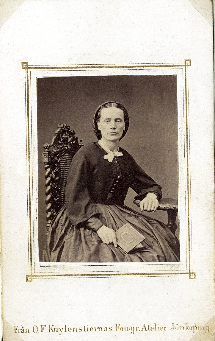 Porträttfoto av en kvinna i mörk krinolin med mörk jacka. Hon sitter på en högryggad stol vid ett bord och håller en liten bok i ena handen.
Knäbild, en face/halvprofil. Ateljéfoto.