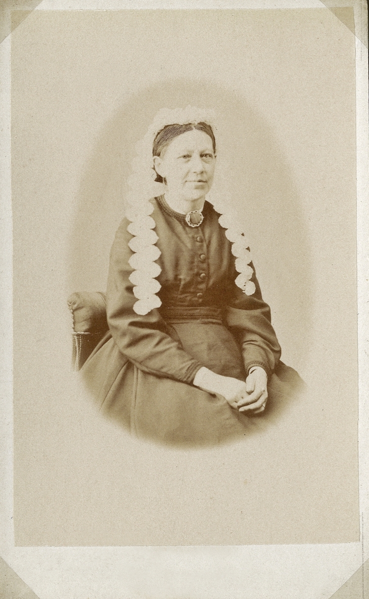 Porträttfoto av en äldre kvinna i mörk klänning och vit hårklädsel/hätta med långa spetsband. Vid kragen syns en stor brosch. 
Knäbild, halvprofil. Ateljéfoto.
