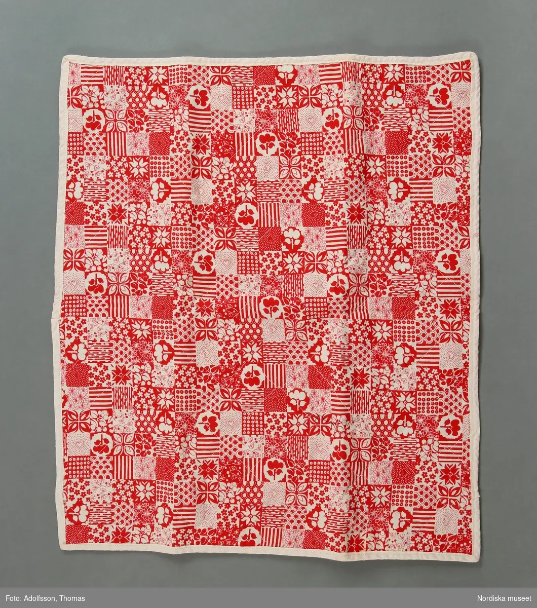 Quiltat maskinsytt docktäcke av bomull och nylon, med tryckt blommigt mönster i rött och vitt. Nylon på undersidan. Kant av vitt bomullsband.