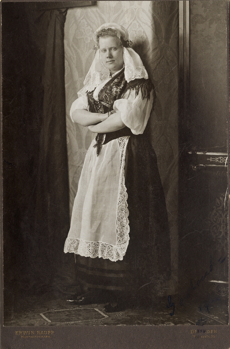 Porträttfoto av en ung kvinna i (trol.) sachsisk folkdräkt. 
I nedre högra hörnet finns en (mycket) otydlig och blekt autograf: "Gertrud, 1903". 
Helfigur. Ateljéfoto.