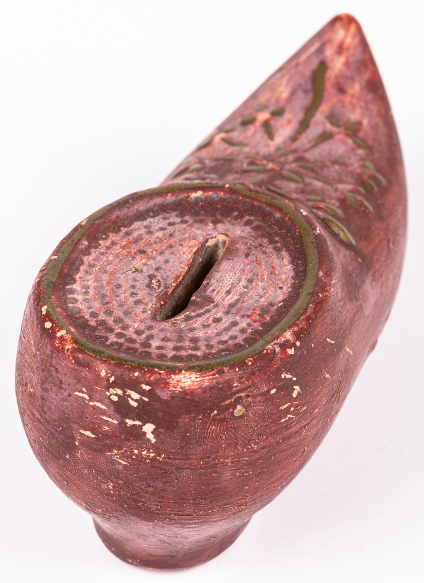 En sparbössa i oljemålat lergods som föreställer en röd träsko/damsko med klack, öppning för mynt och dekorerad med blommönster upptill i guldbronsering. Innuti ligger en liten knapp.